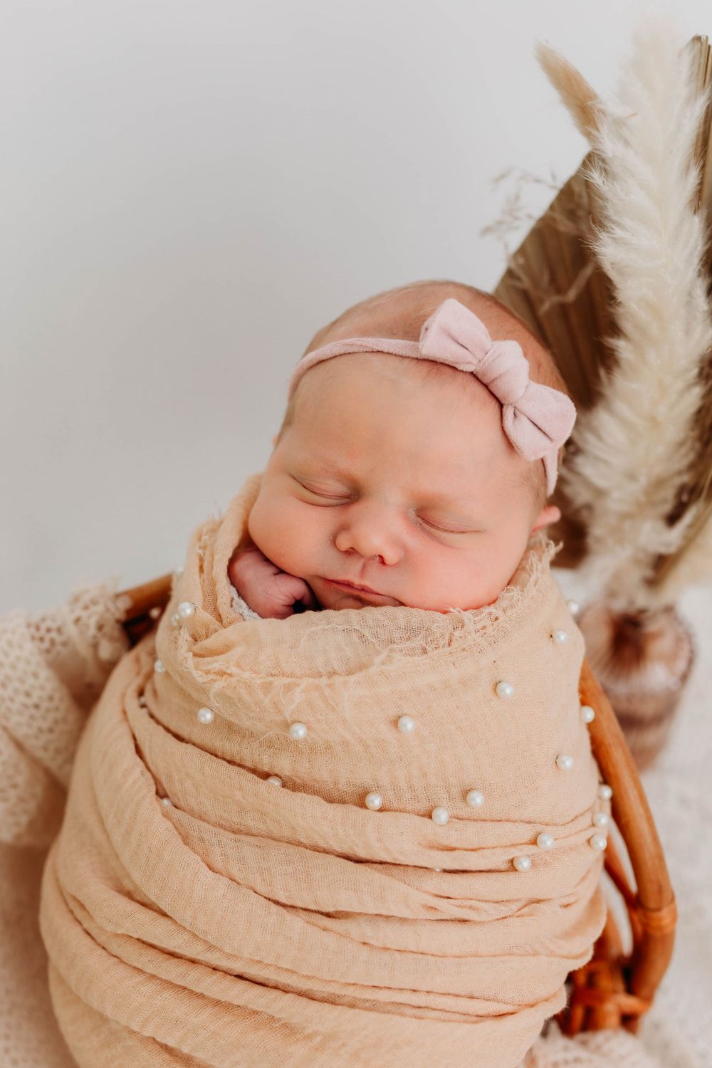 Sleeping newborn swaddled in beige wrap with pink bow headband and feather decor.