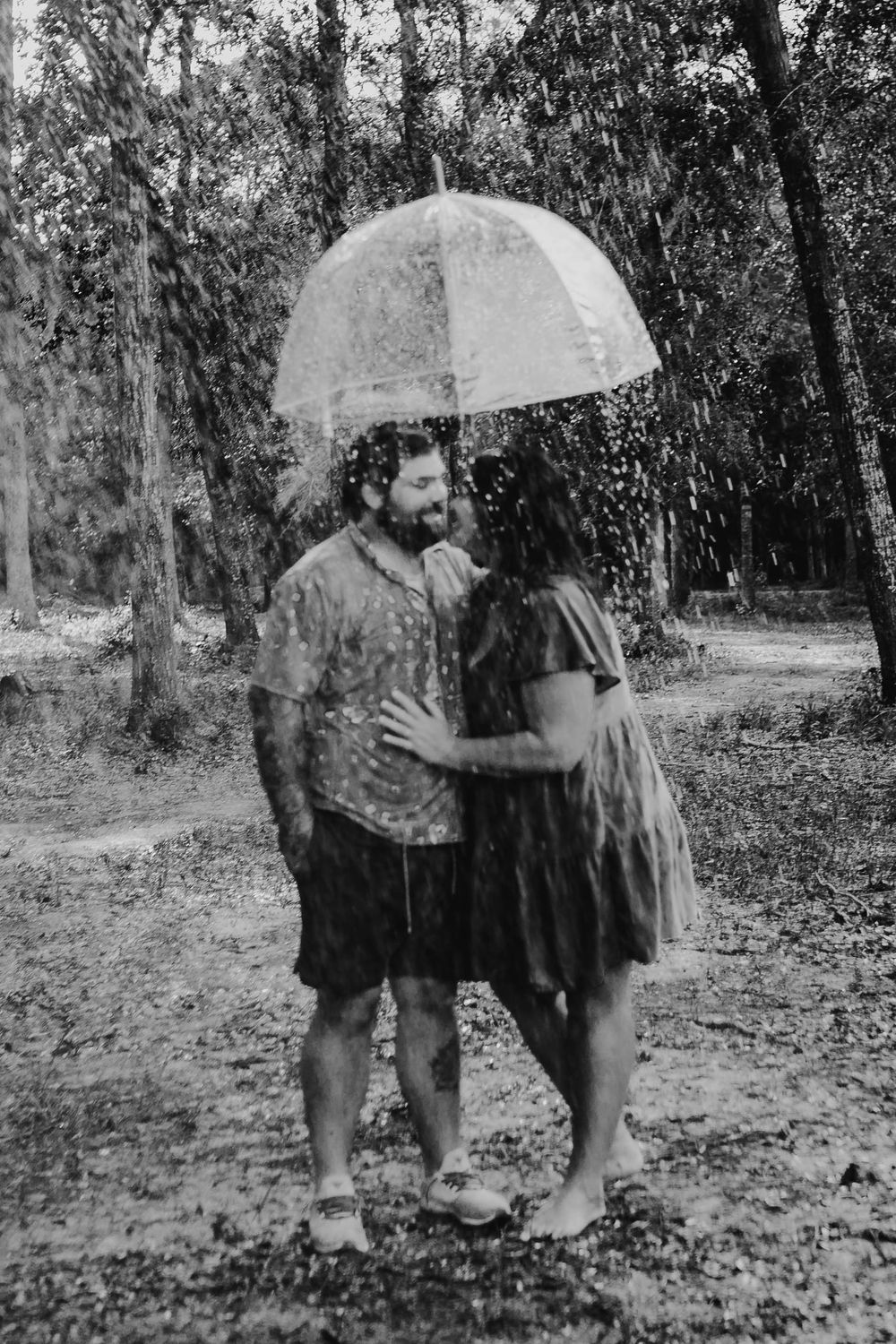 A romantic couple sharing an umbrella in the rain within a forest setting in black and white.