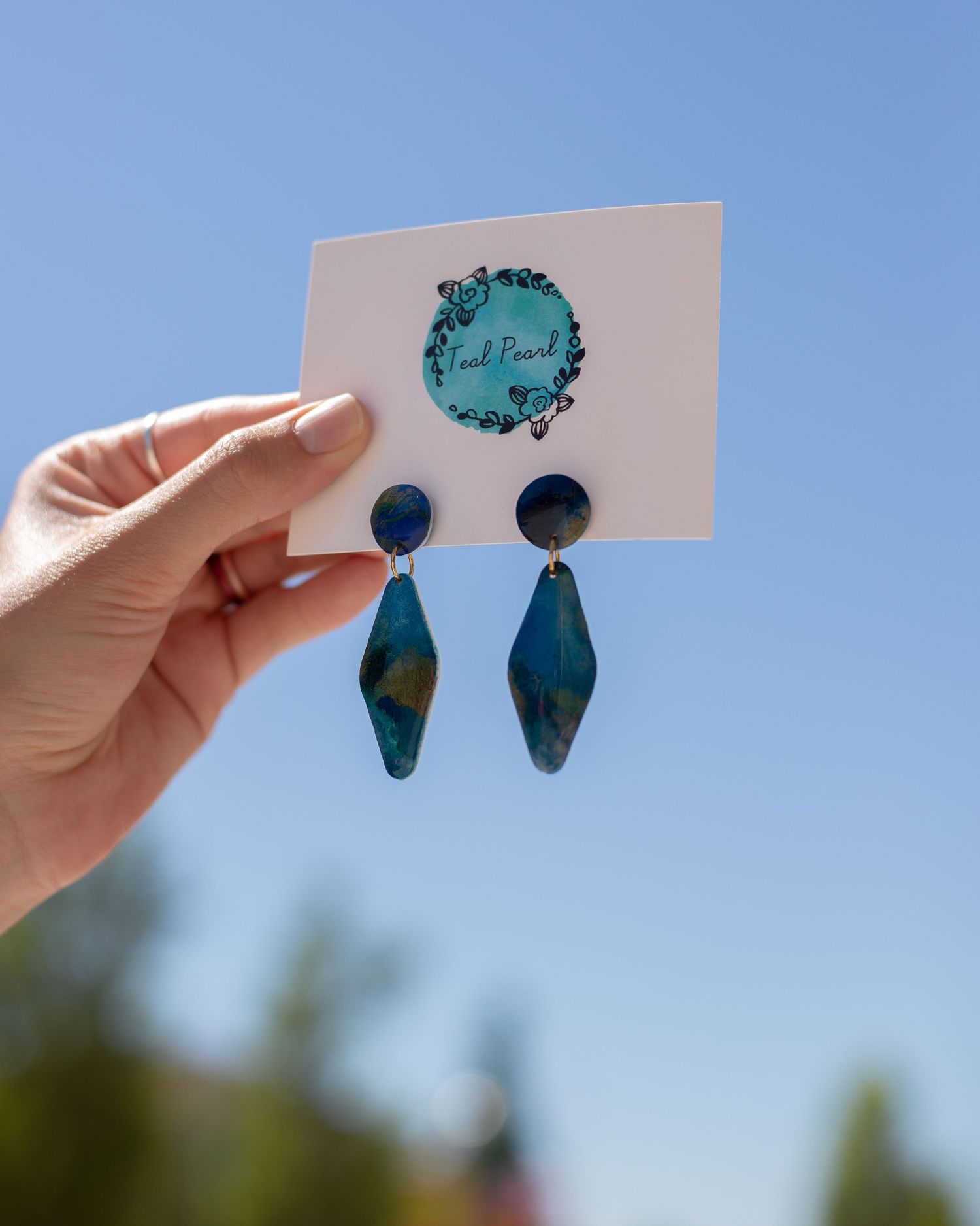 A hand holds up a white card mounted with iridescent blue earrings made with polymer clay and a clear coat of resin. Blue sky in background.
