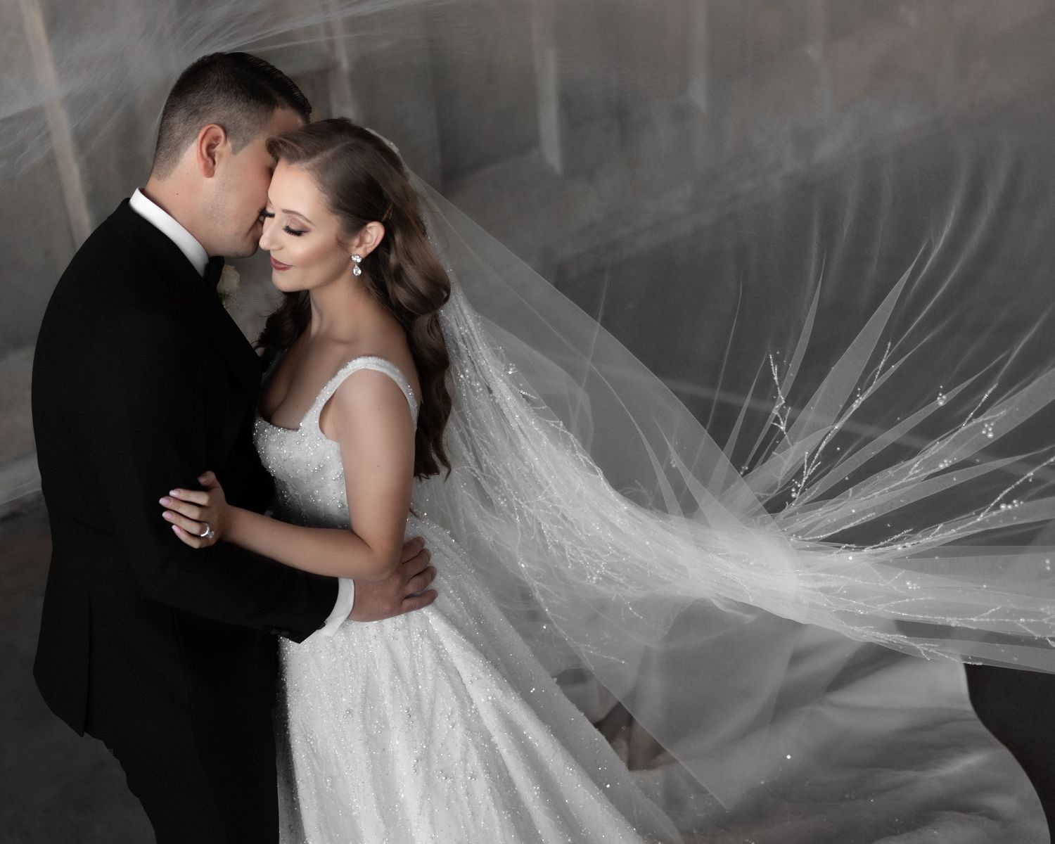 Bride in lace gown with flowing veil embraces groom in black tuxedo in dramatic black and white photo.