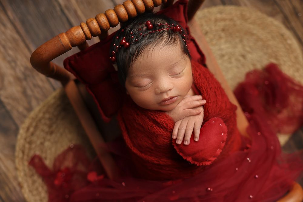 Newborn baby sleeps peacefully wrapped in red fabric on wooden prop with delicate beaded details.