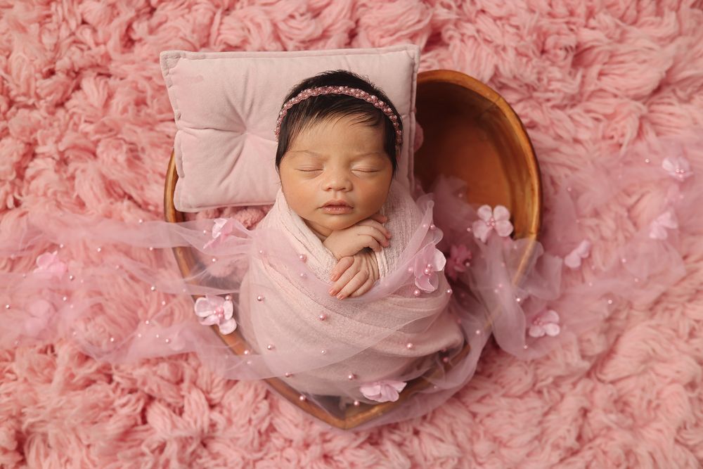 Sleeping newborn wearing pink tulle dress and headband lies in wooden bowl on fluffy pink backdrop.