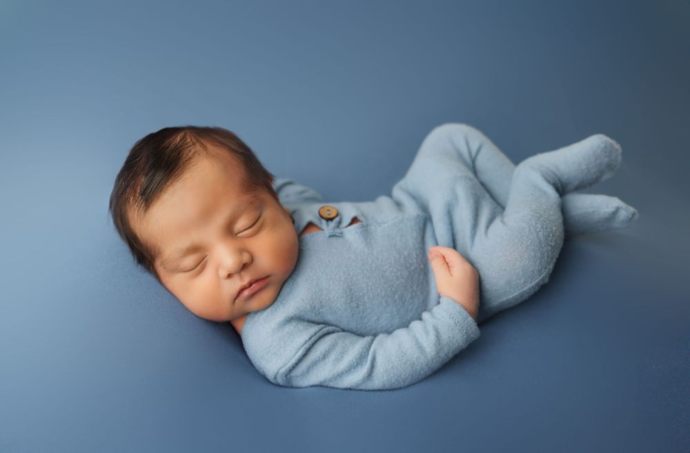 Peaceful sleeping newborn wearing soft blue sleeper outfit lying on blue backdrop during professional photo session.