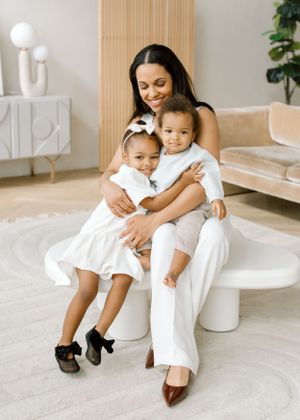 Parent and two children in white outfits share loving embrace on white stool.