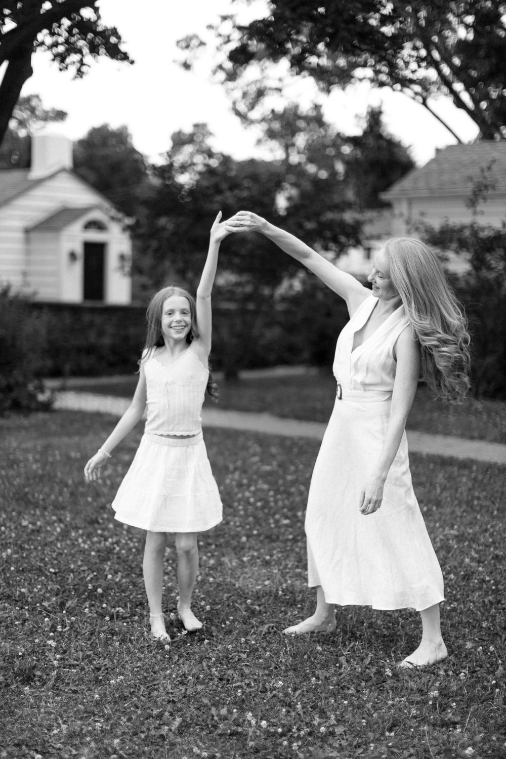 Two figures in white dresses dance together joyfully in a backyard with houses visible in the background.