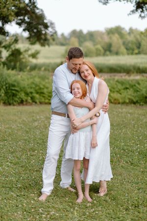 A family dressed in white outfits embraces while standing together in a sunlit grassy field during a summer photo session.