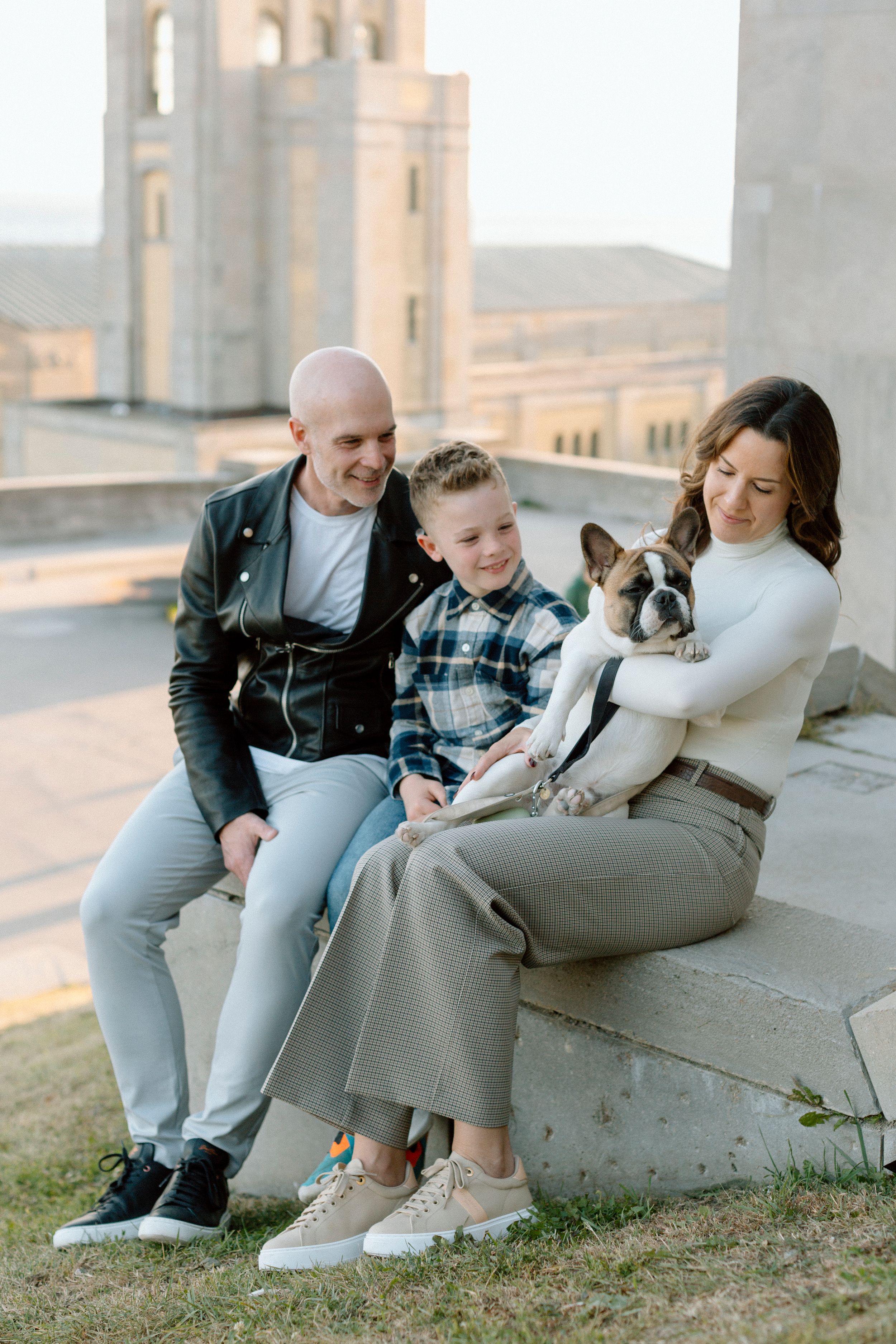 Family sits together on concrete steps with their French Bulldog while visiting a cathedral during golden hour.
