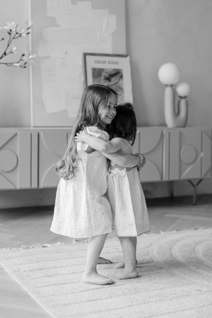 Two small children in white dresses share a tender embrace while standing barefoot on a rug in a monochrome scene.