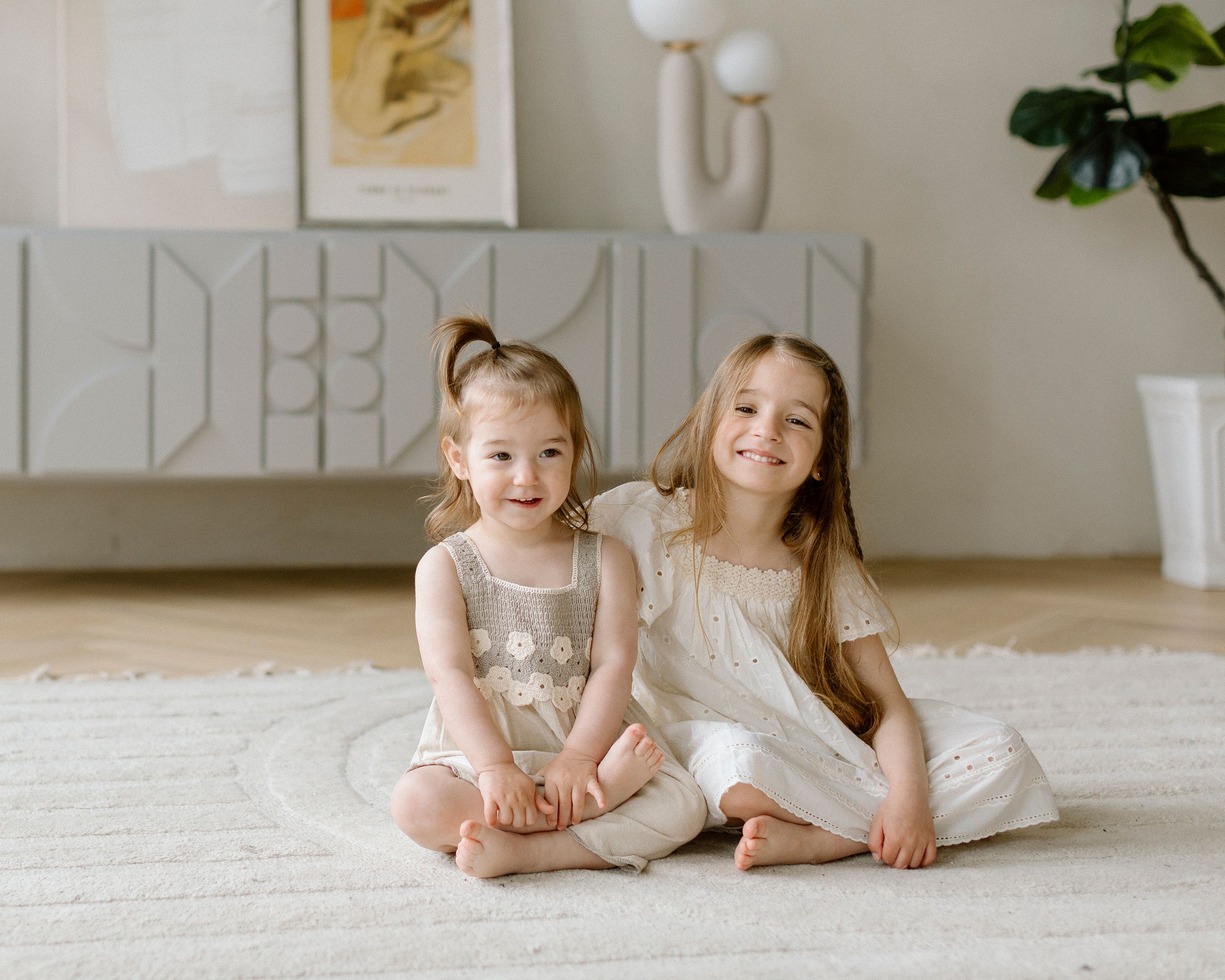 Two children sitting together on a white carpet wearing light colored dresses in a minimal modern living room.