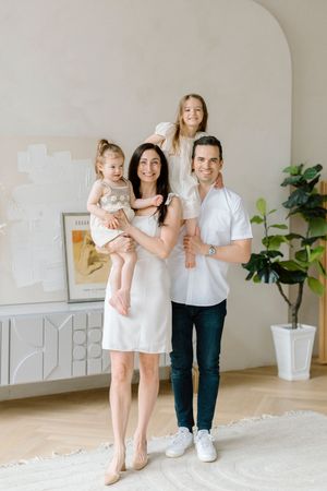Family poses together in front of a light colored wall with a decorative frame and house plant in a modern interior setting.