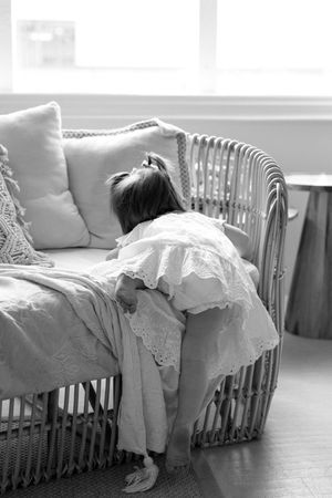A small figure in a white lace dress reclines on a wicker couch with cushions near a window in black and white.