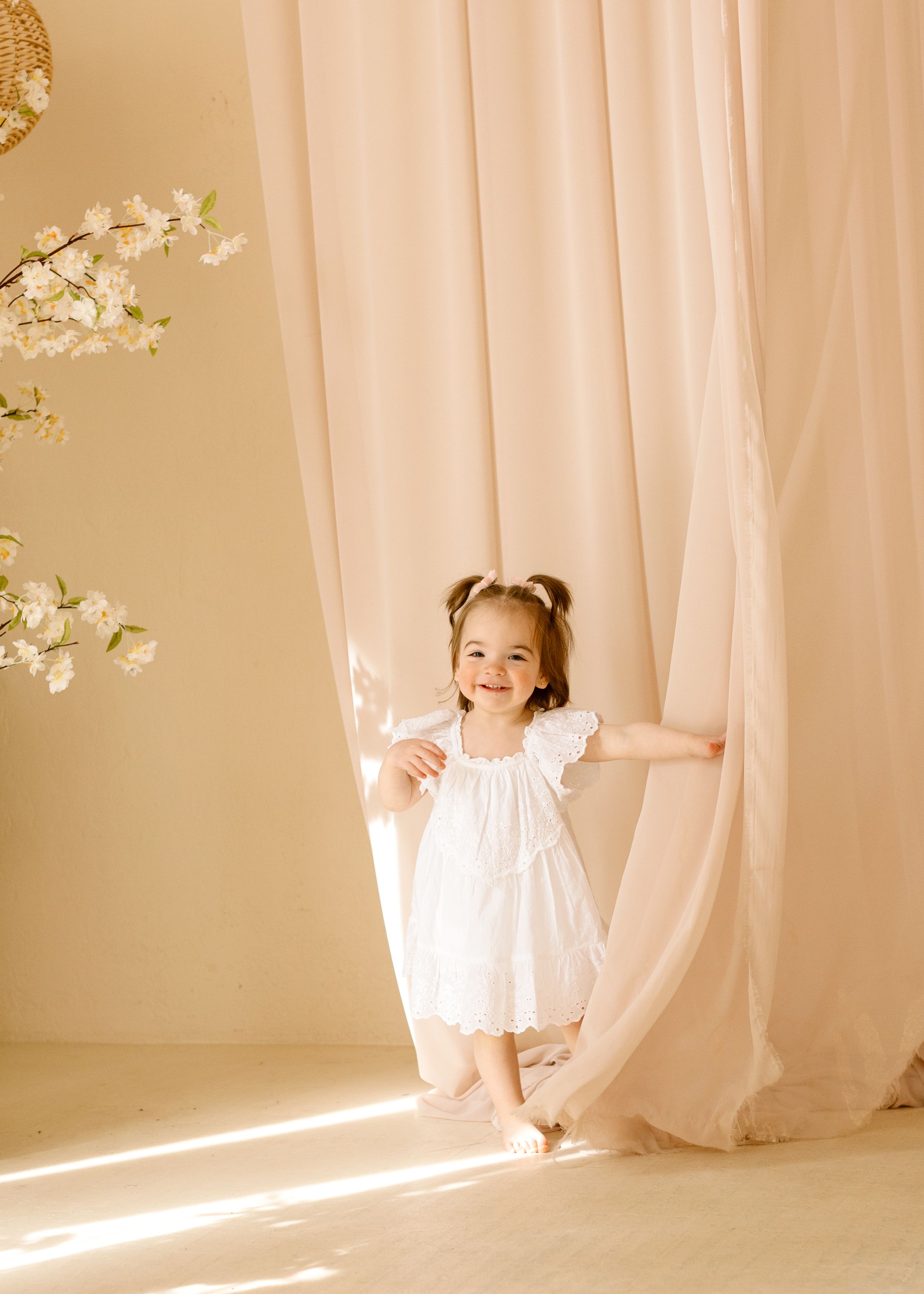 A young child in a white dress dances and plays with flowing curtains in a softly lit room filled with natural sunlight.