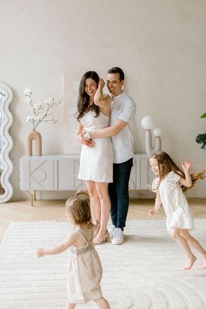 A family shares a tender moment in a bright minimalist room while two young children dance playfully on a white rug.