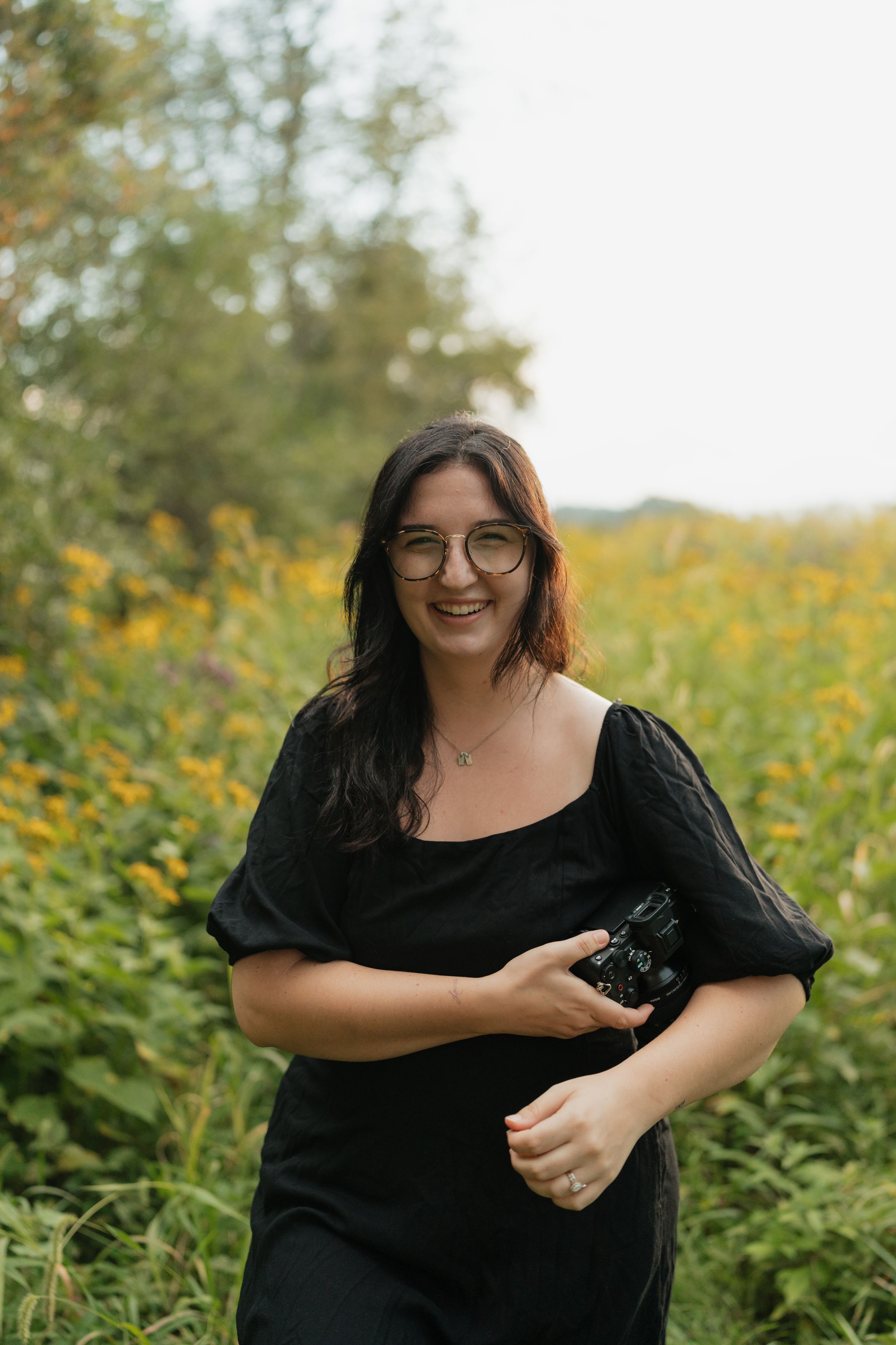 A person in black holding a camera poses in a field of yellow wildflowers.