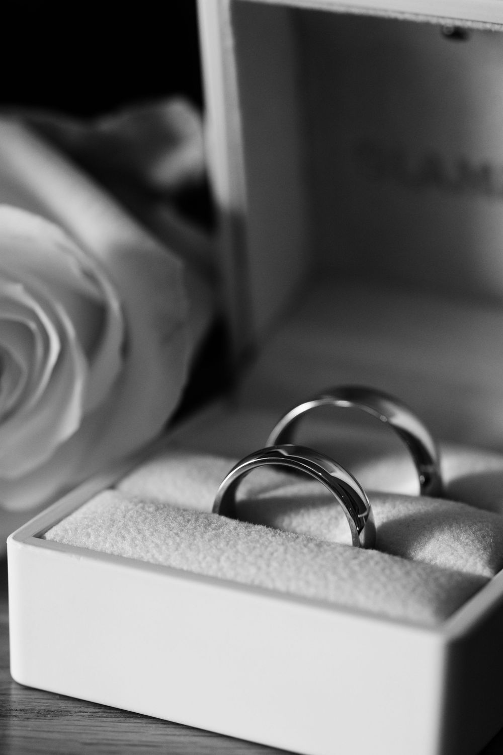 Close-up black and white photo of two wedding rings nestled in a white ring box with a soft rose in the background.