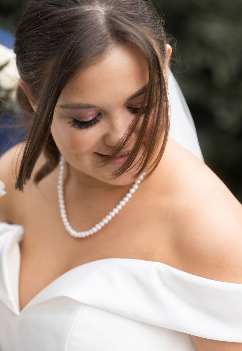 A bride in a white wedding dress and pearl necklace looks down with a gentle smile in a soft-focused portrait.