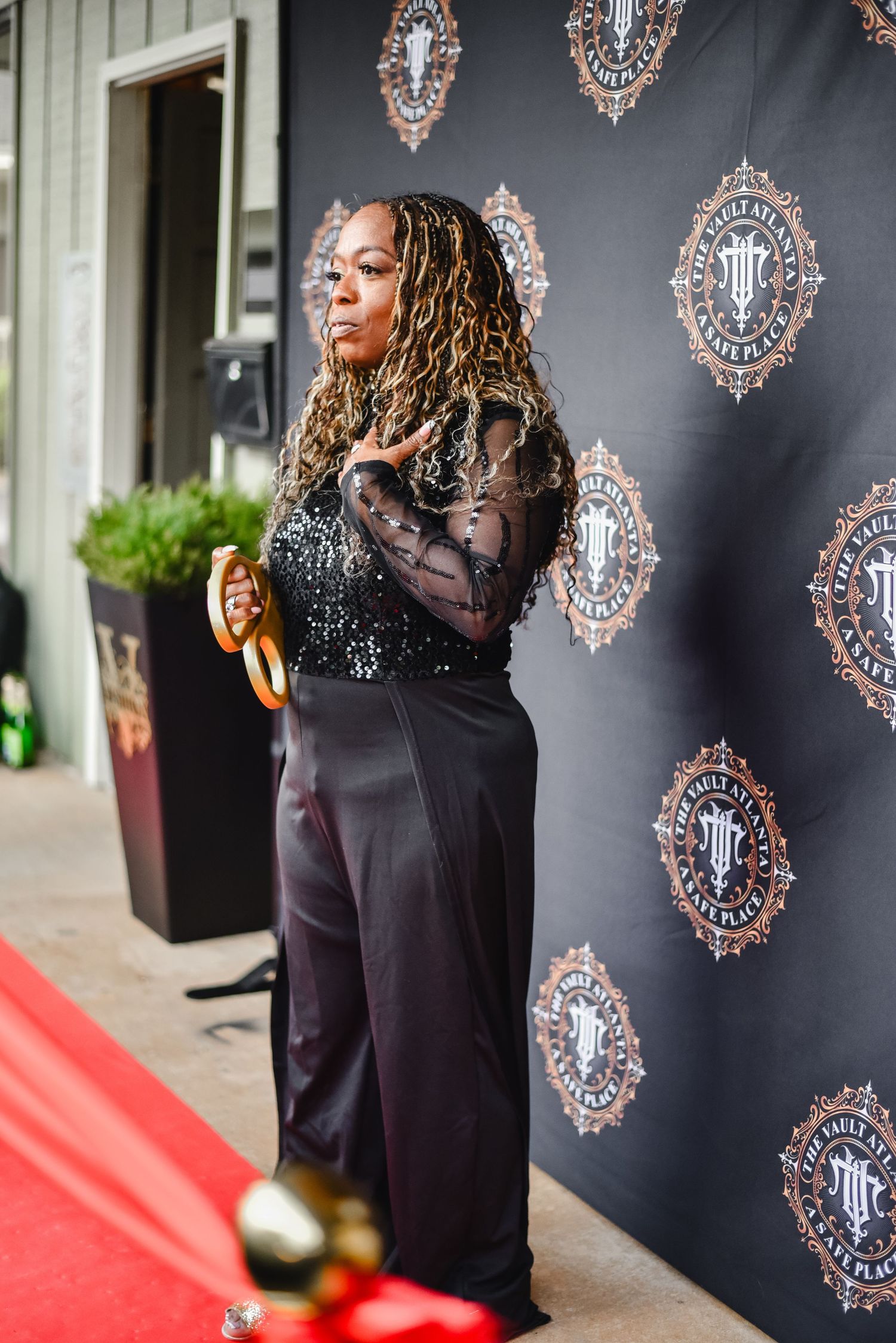 Glamorous figure poses on red carpet against black backdrop with ornate logo designs.