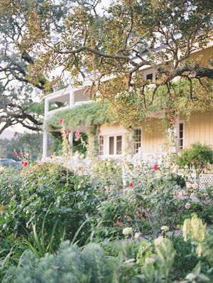 A charming country house with white columns and porch surrounded by a lush garden with blooming flowers and trees. Wedding at Beltane Ranch