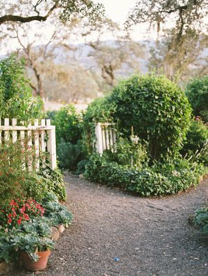 A winding garden path bordered by lush greenery and white picket fencing leads through a tranquil outdoor space. Cottage garden wedding