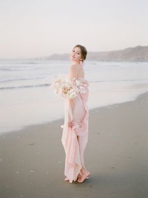 A bride in a flowing pink gown walks gracefully along a misty beach at sunset, creating a romantic coastal scene. Beach wedding in Bay Area