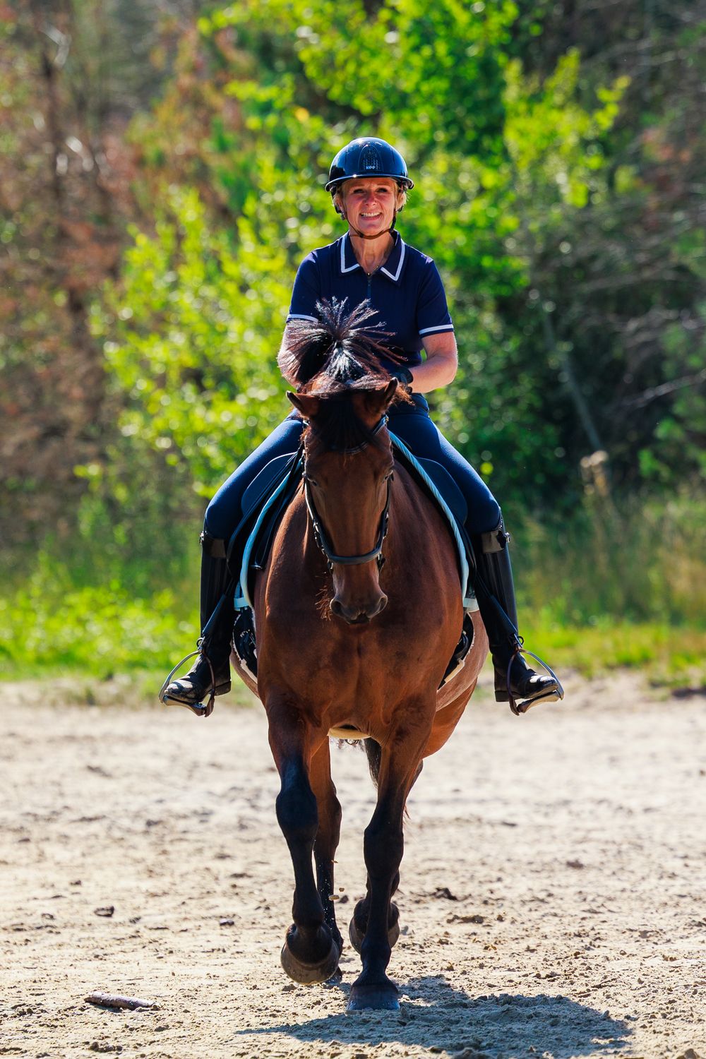 A horse and rider perform a galloping sequence on a sandy riding arena with green trees in the background.