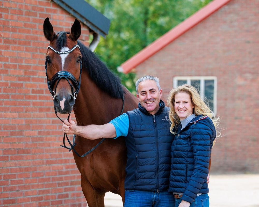 A happy couple poses outdoors with their beautiful bay horse against a brick building backdrop.