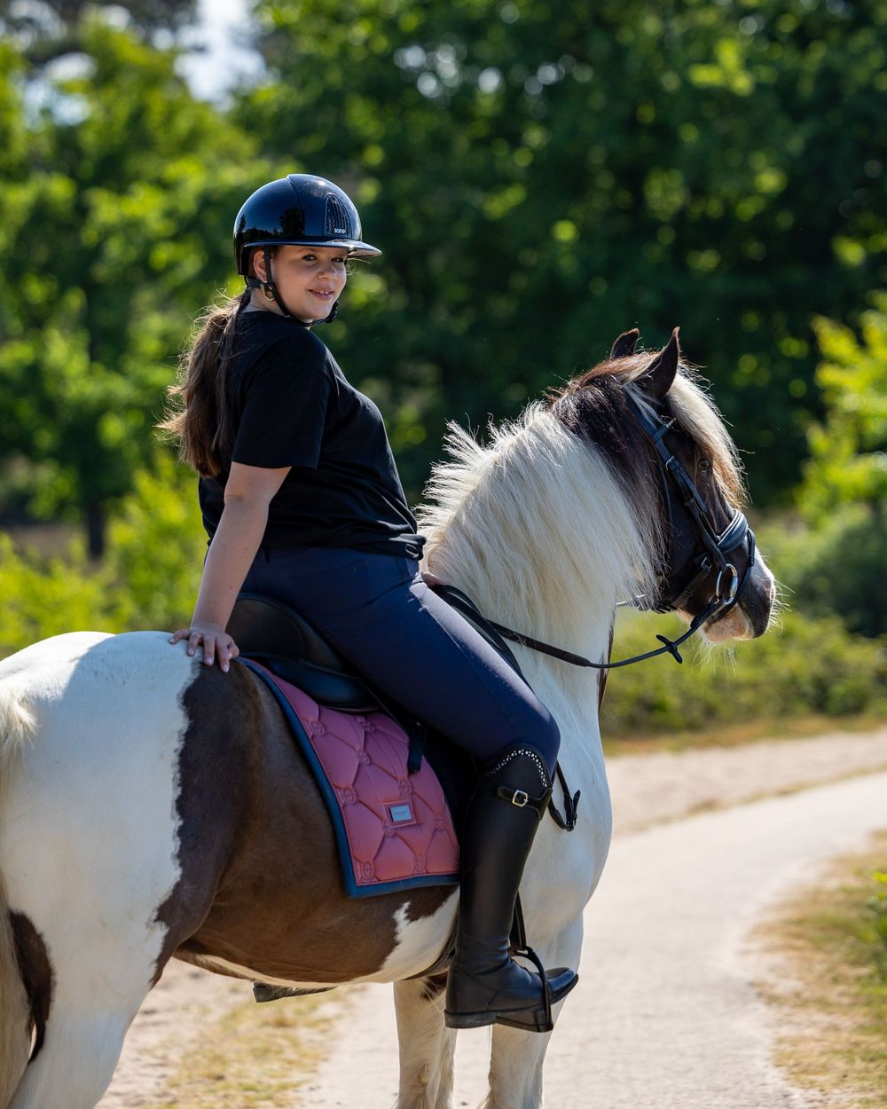 An equestrian in a helmet rides a black and white horse on a sunlit dirt path during a summer day.