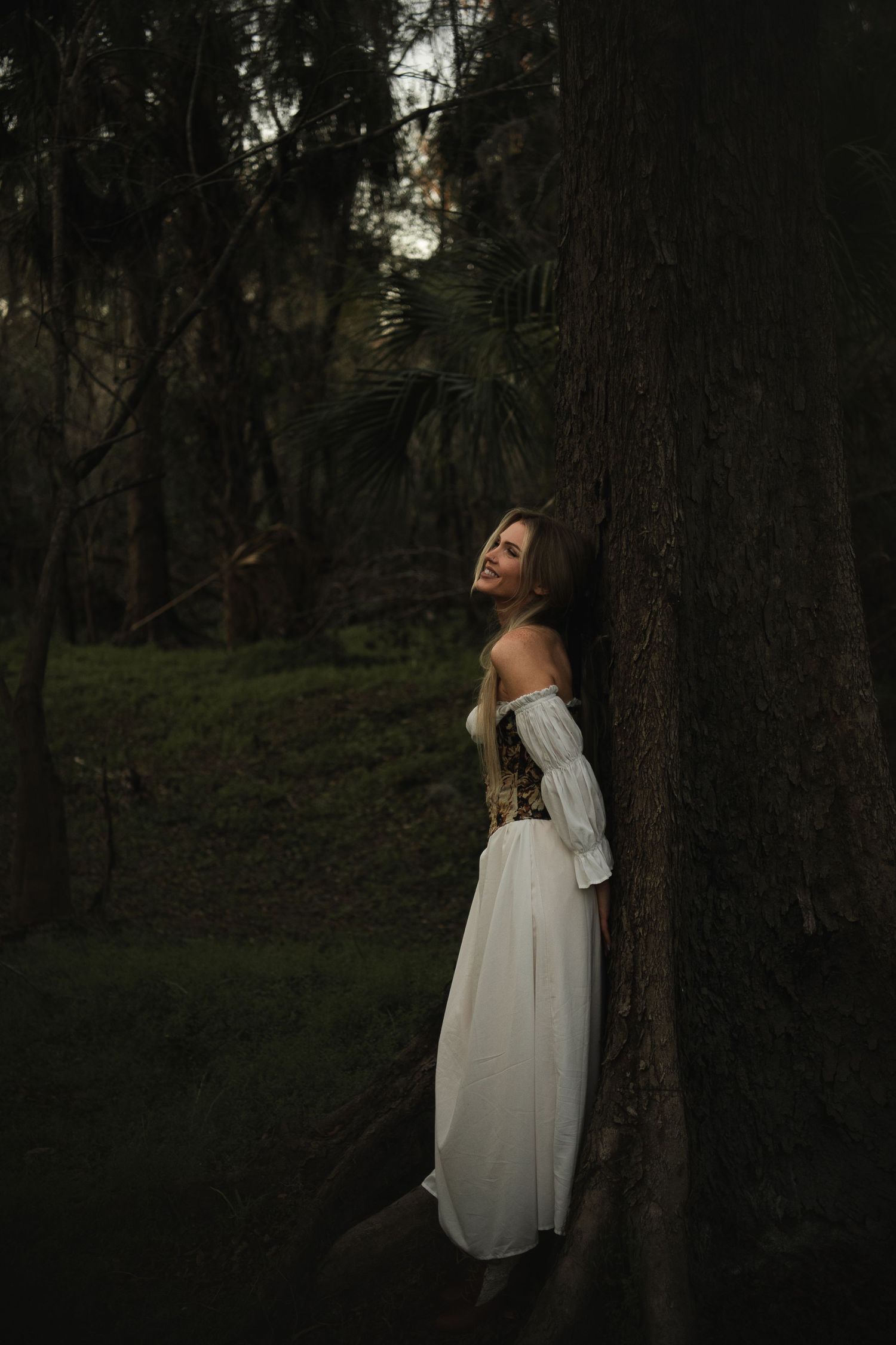 Woman in white off-shoulder dress leaning against large tree trunk in dark forest setting.