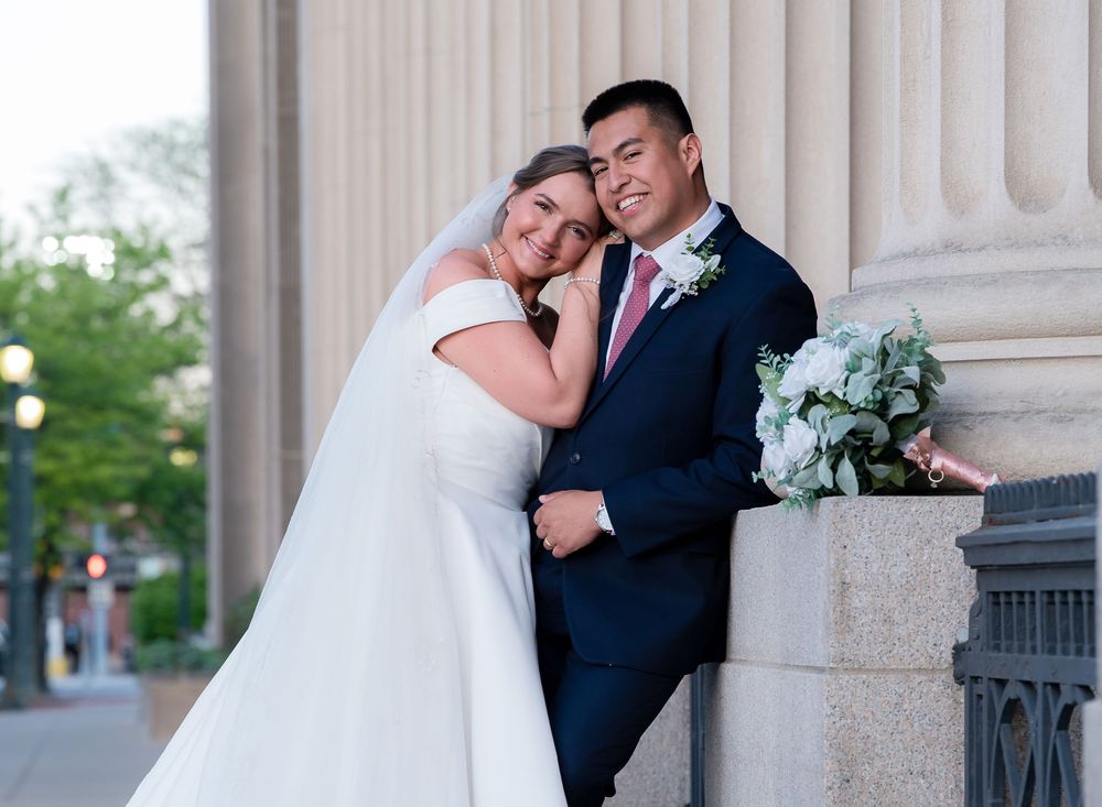 A romantic wedding couple poses together against classic white stone columns in an elegant outdoor setting.