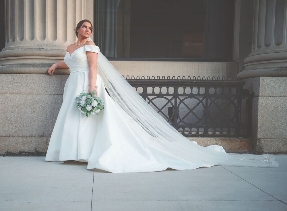 A bride in a flowing white wedding gown poses elegantly against a classical stone column backdrop.