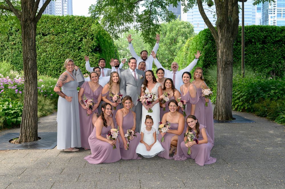 A wedding party in lavender dresses poses together in a lush garden setting with tall trees in the background.
