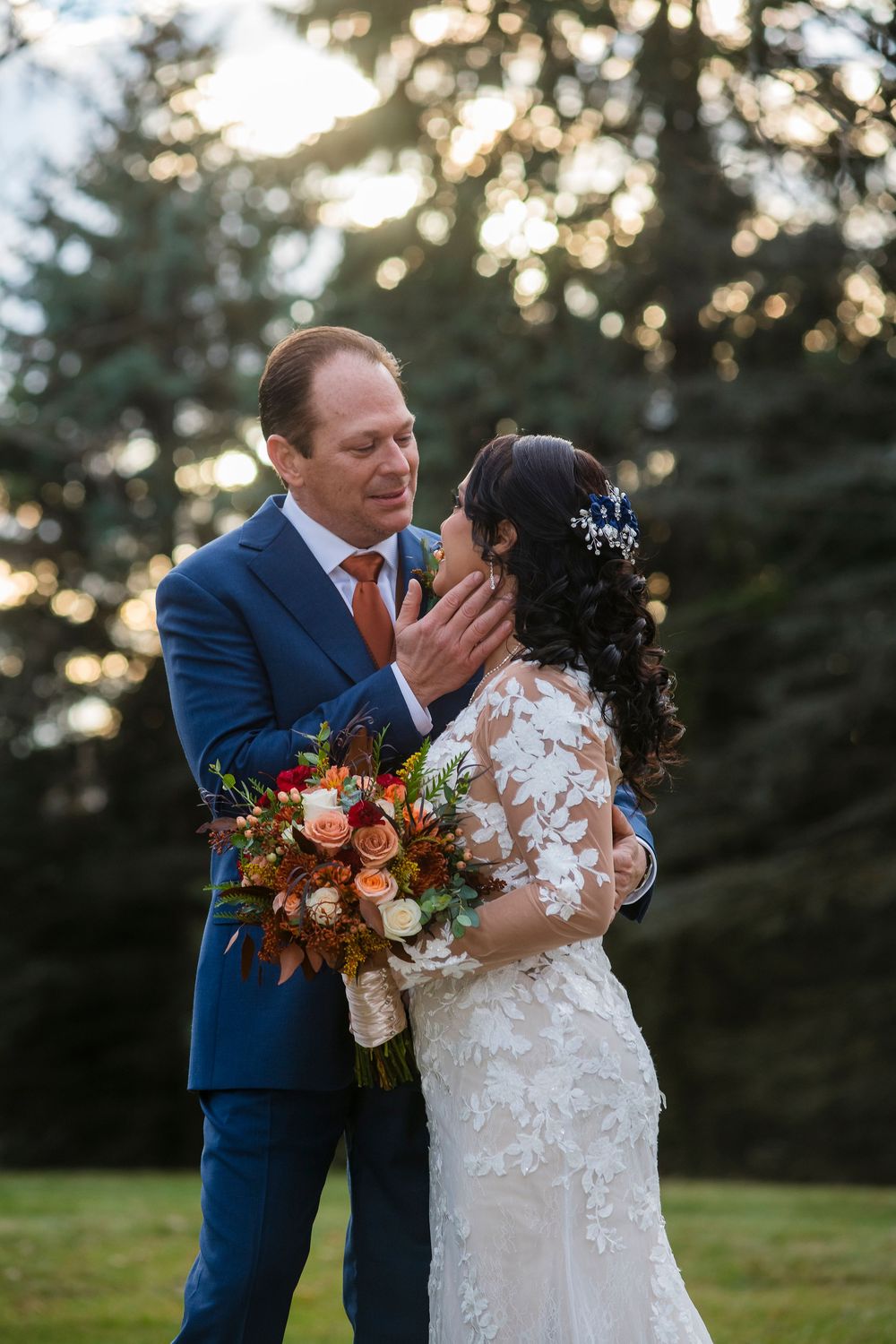 A romantic wedding couple embraces in a golden sunset with autumn florals and a navy suit against an evergreen backdrop.
