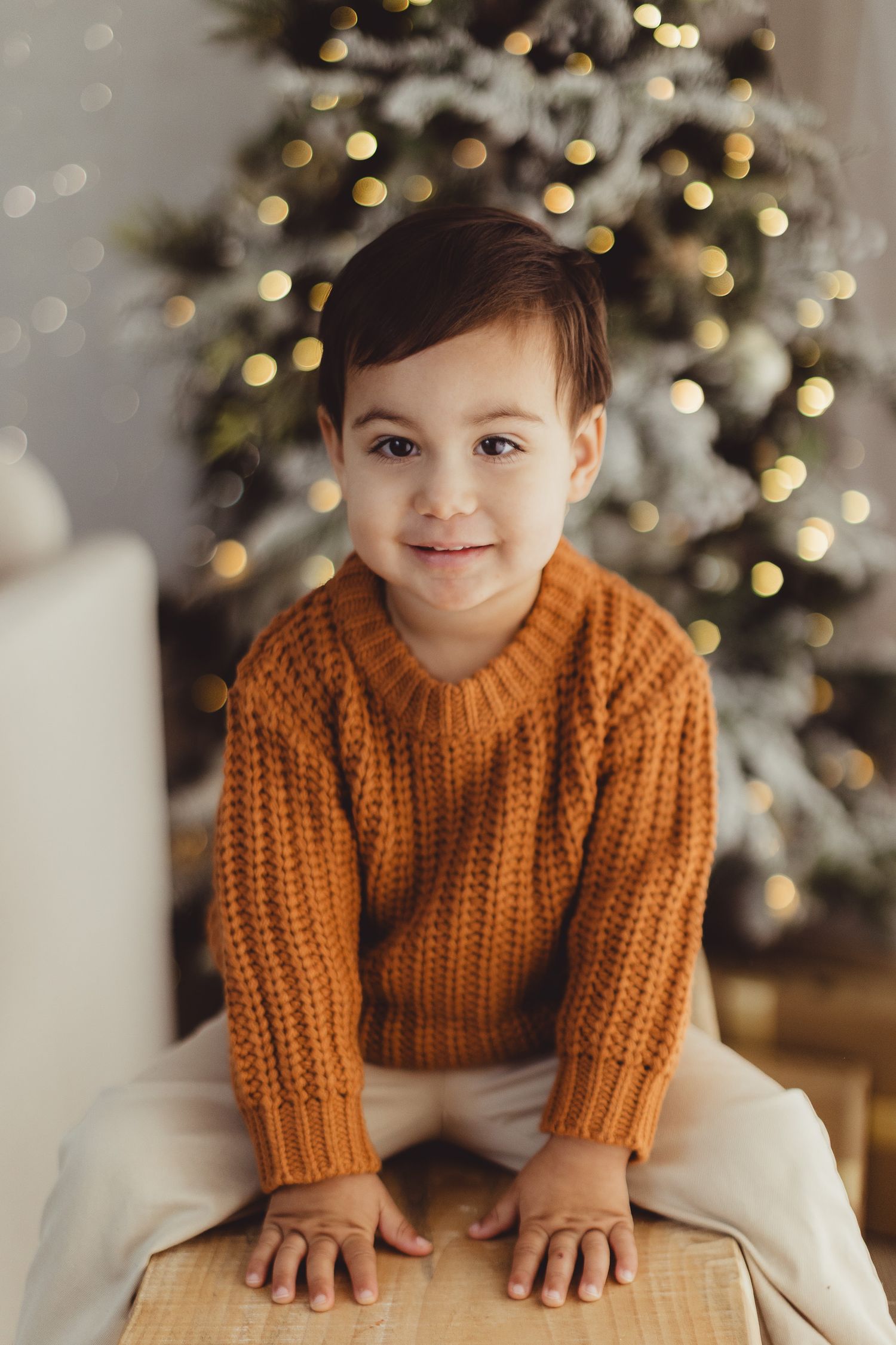 Child in orange knit sweater poses playfully on white chair with Christmas tree backdrop.