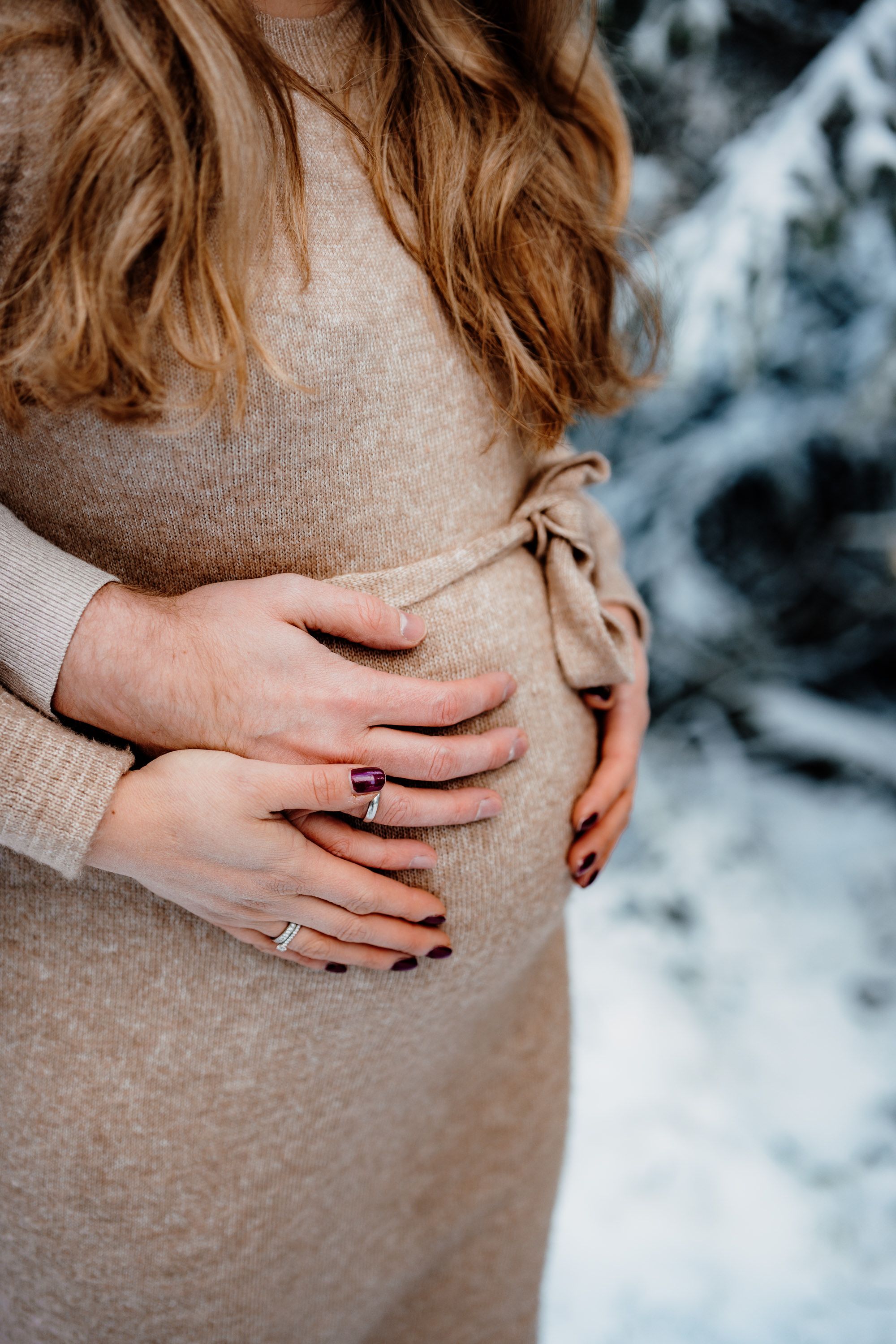 Close-up of hands resting on a pregnant belly wearing a beige dress in a snowy setting.