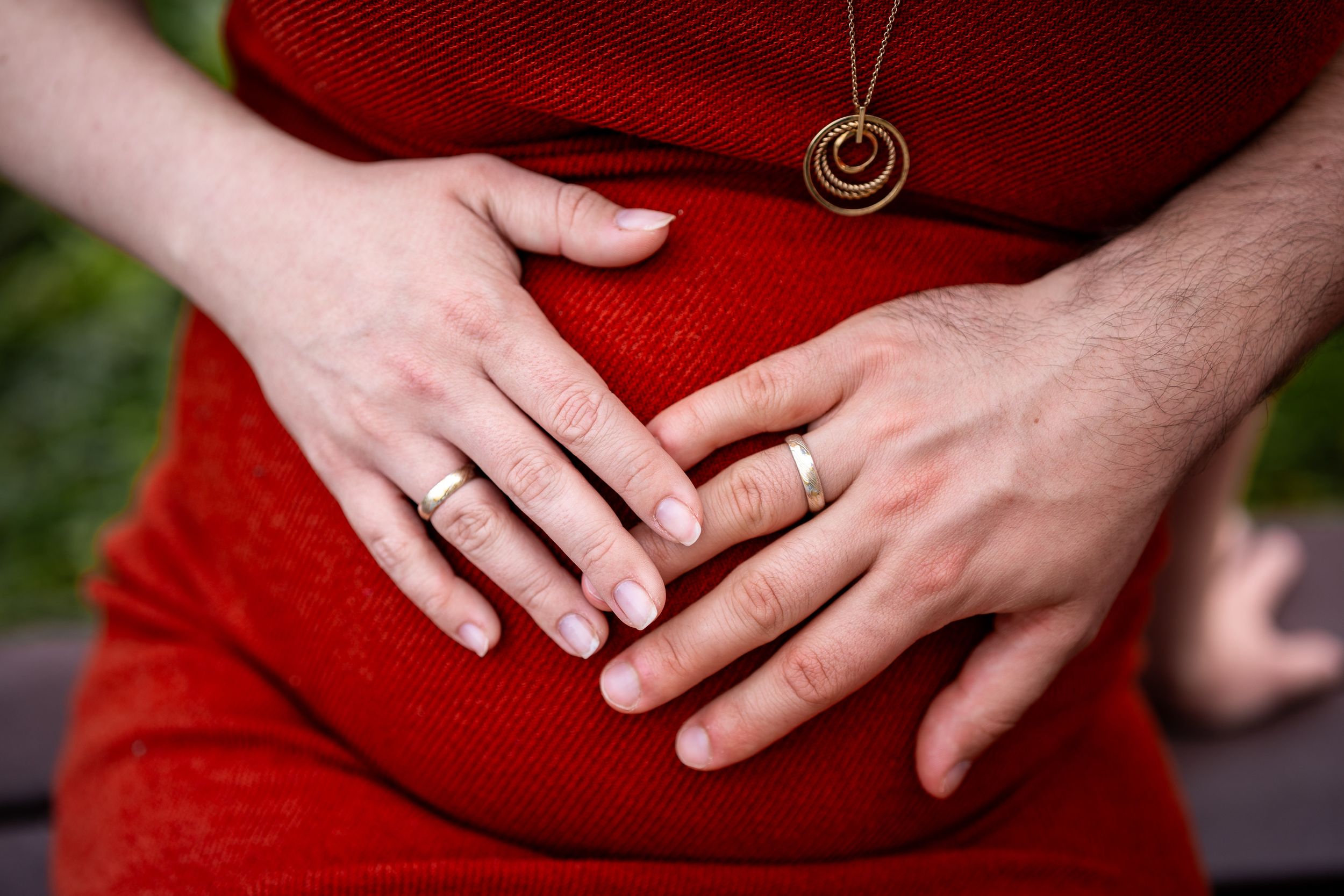 Close-up of hands with wedding rings resting on red dress with necklace detail.
