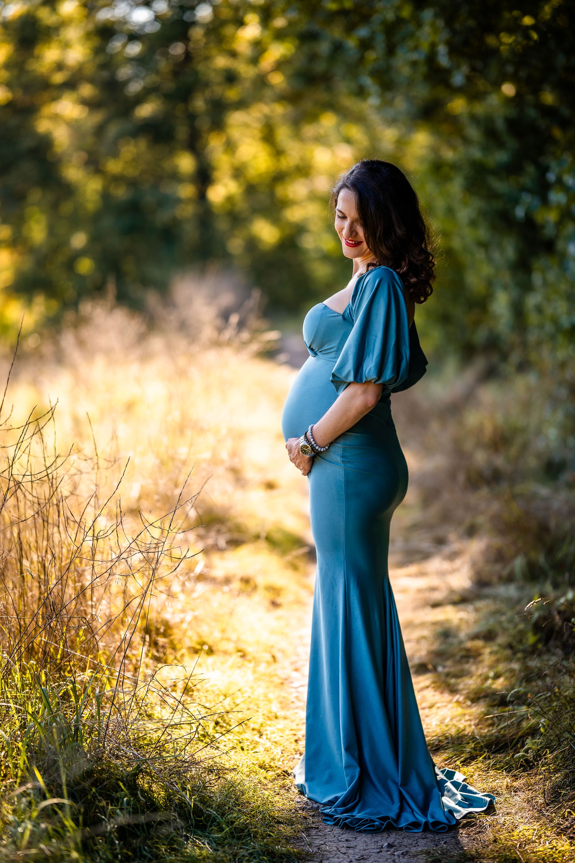 Maternity photo of expectant mother in flowing blue dress against golden sunset background.