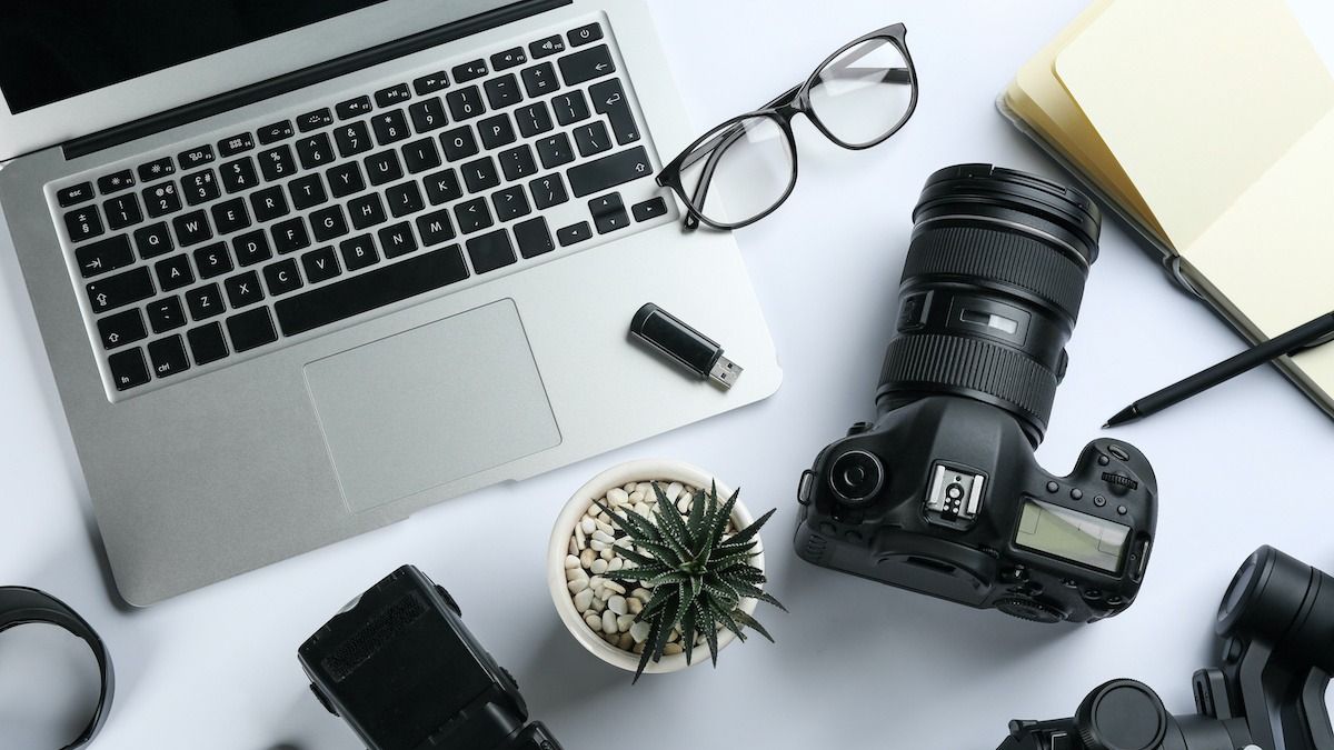 Flat lay of workspace with laptop, camera equipment, glasses, and small potted plant on white desktop.