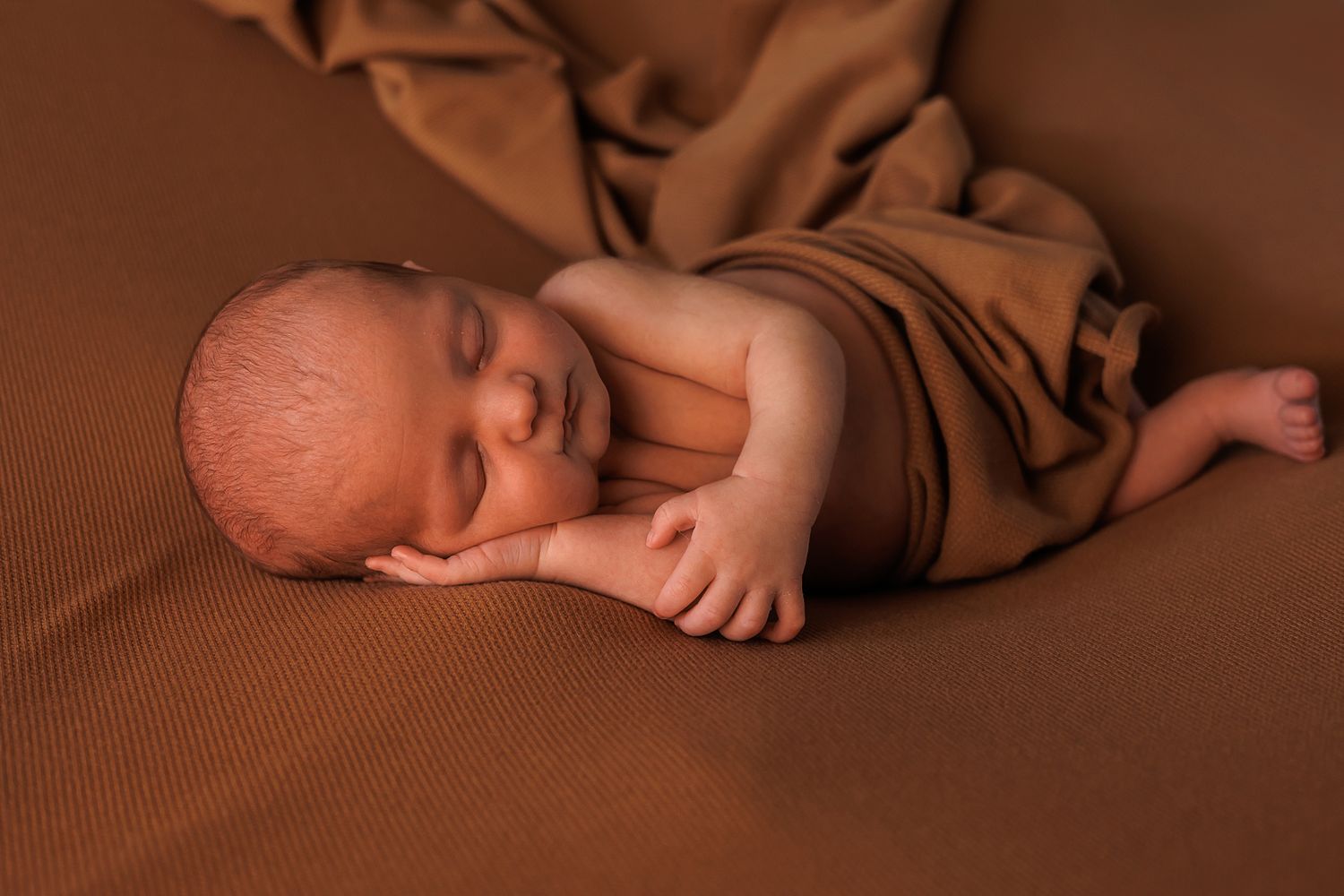 Newborn baby sleeping peacefully on brown fabric backdrop.