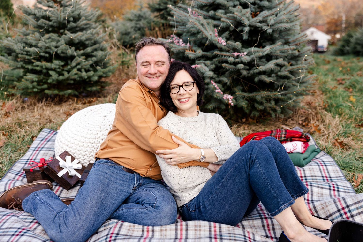 Family relaxing together on a plaid blanket with evergreen trees in the background.