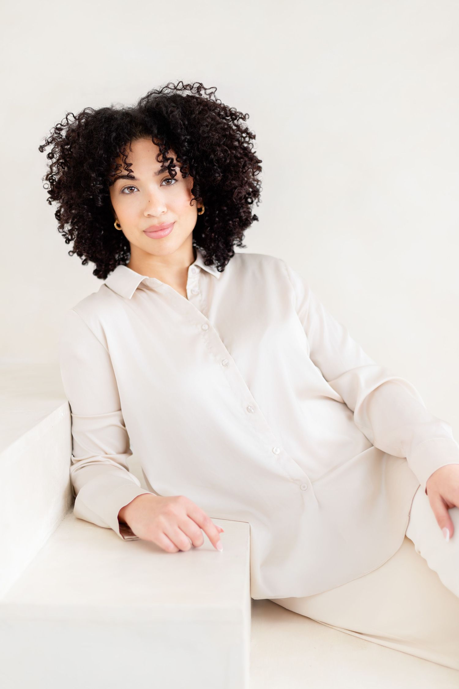 Person with curly dark hair wearing a white button-down shirt poses casually on light colored furniture.
