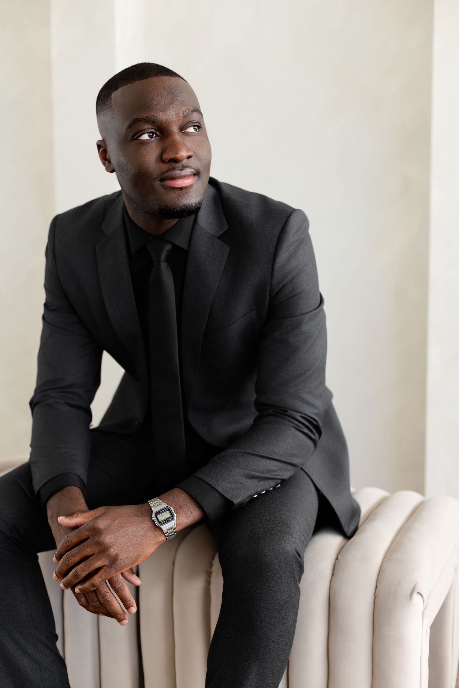 Professional portrait of someone in a sleek black suit and tie sitting casually on a white radiator with a warm smile.