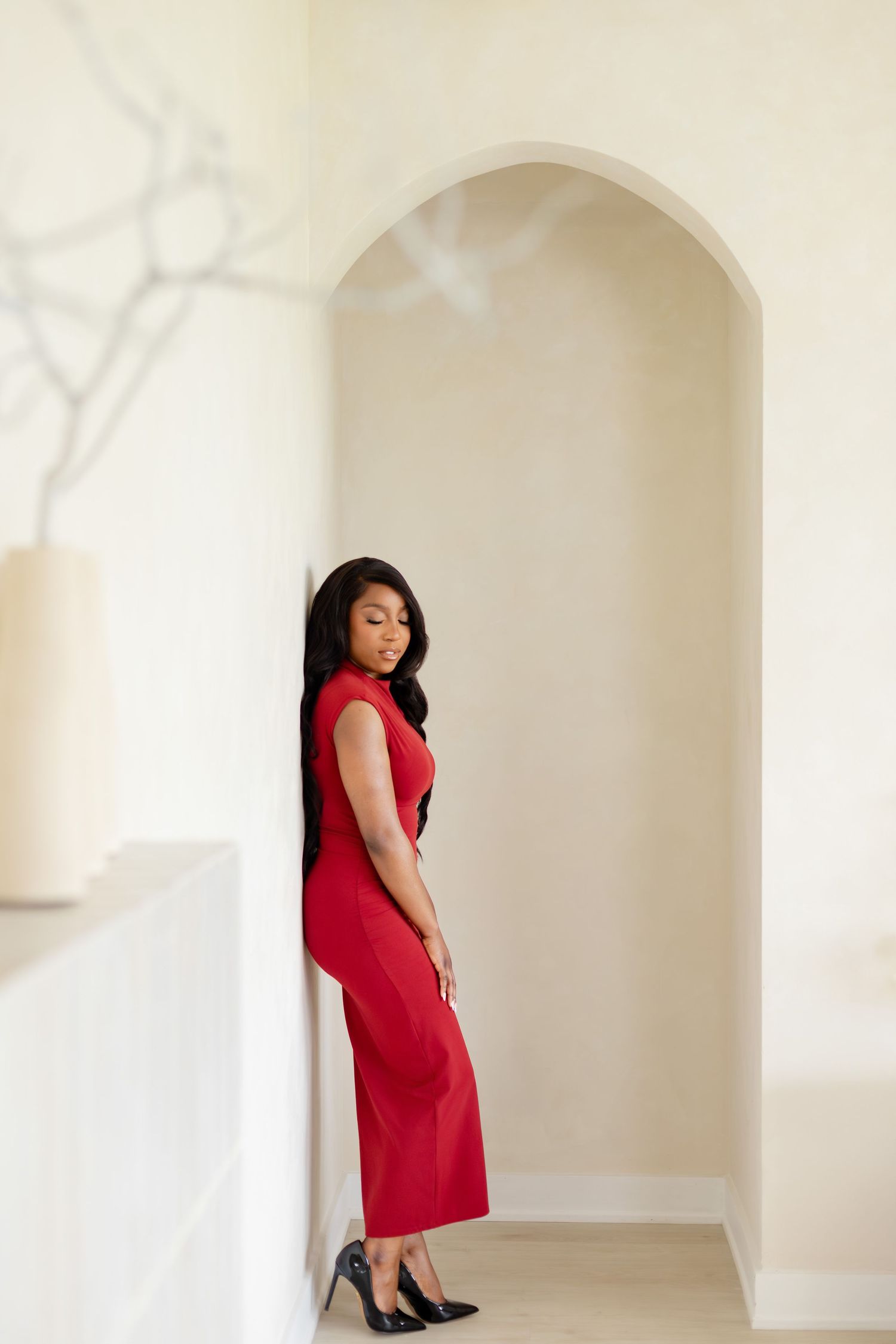A woman in a red sleeveless dress and black heels poses confidently in an arched white hallway alcove.