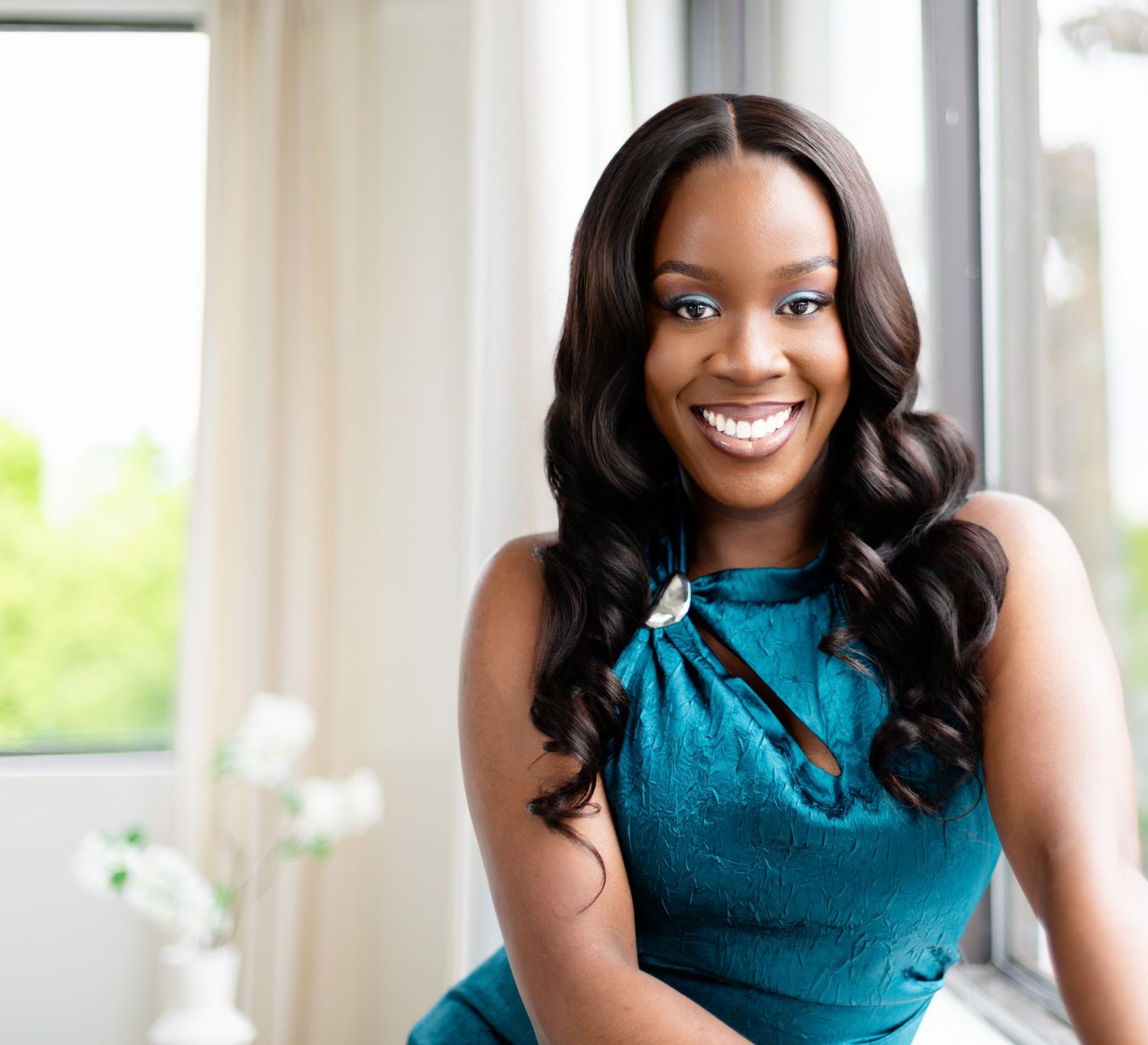 A person in a teal sleeveless top poses against a bright window backdrop while smiling naturally.
