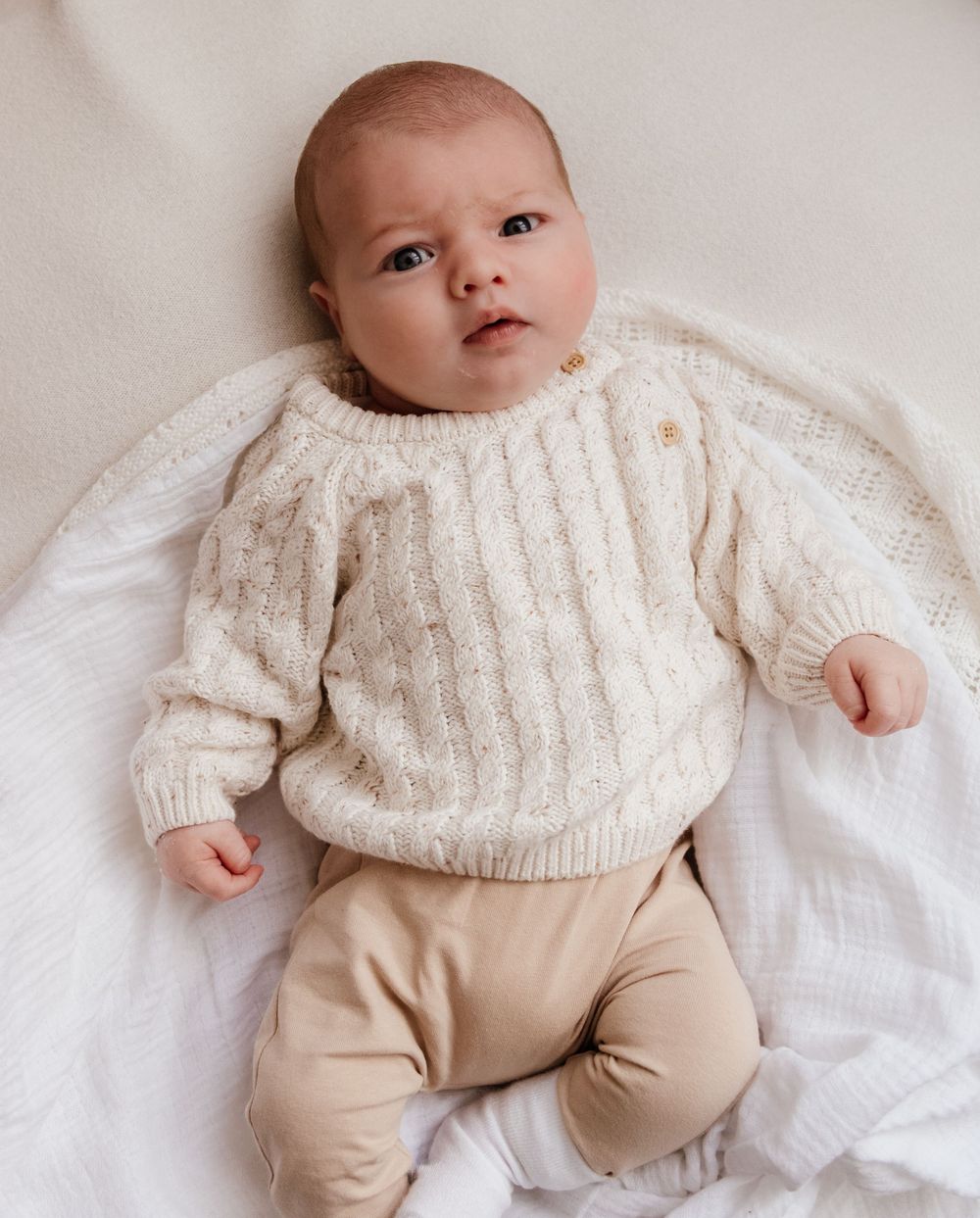 Baby wearing cream cable knit sweater lying on white bedding in lifestyle portraits.