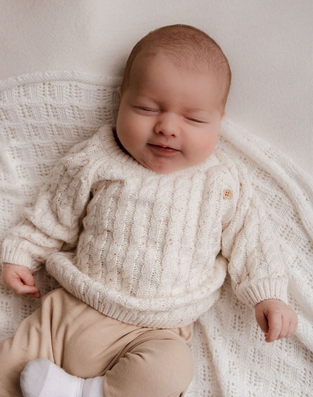 A sleeping baby wearing a cozy white knit sweater and beige pants lies peacefully on a light-colored backdrop.