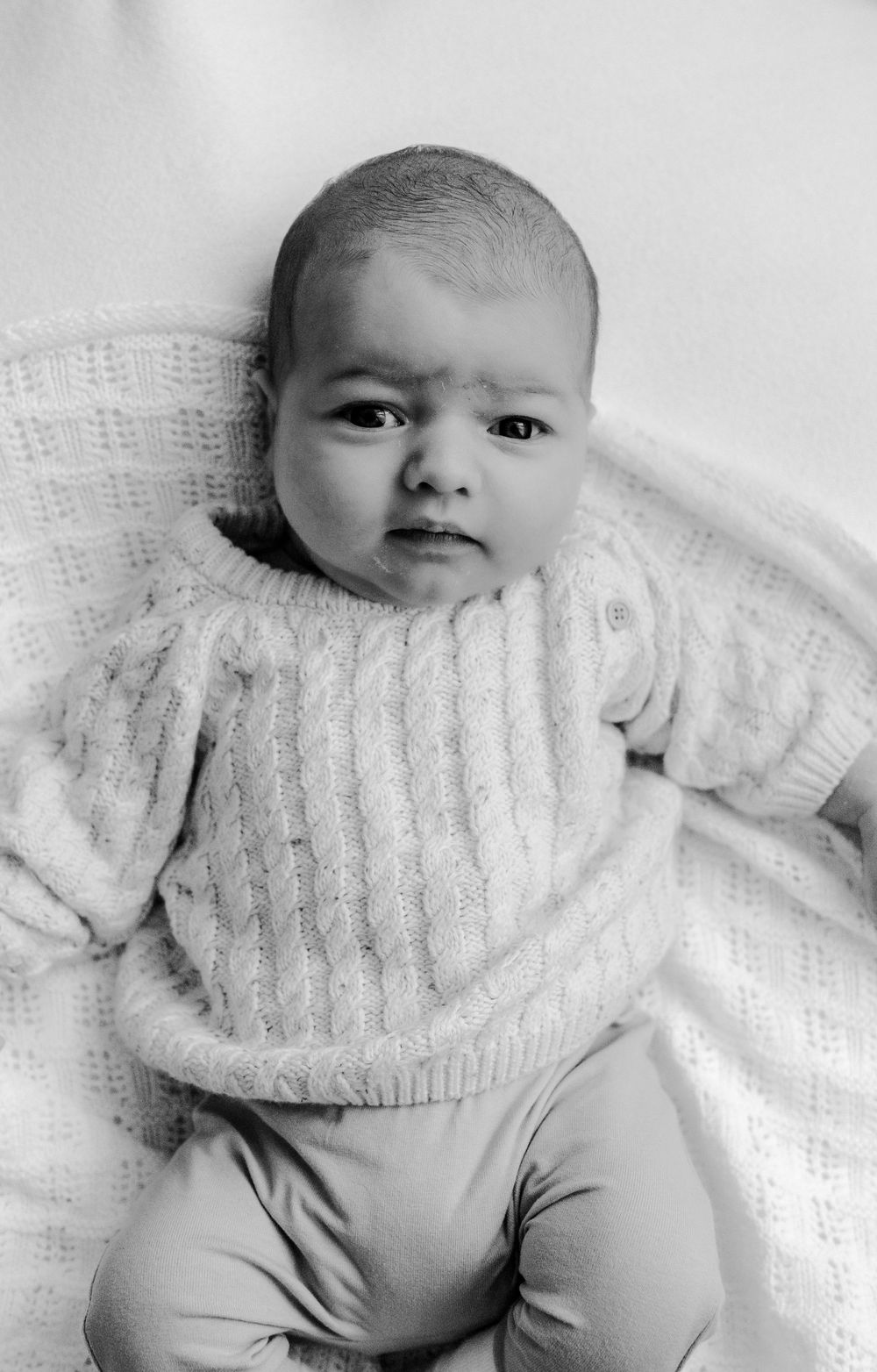 A black and white photo series shows a baby wearing a cozy white knit sweater while lying on a light background.