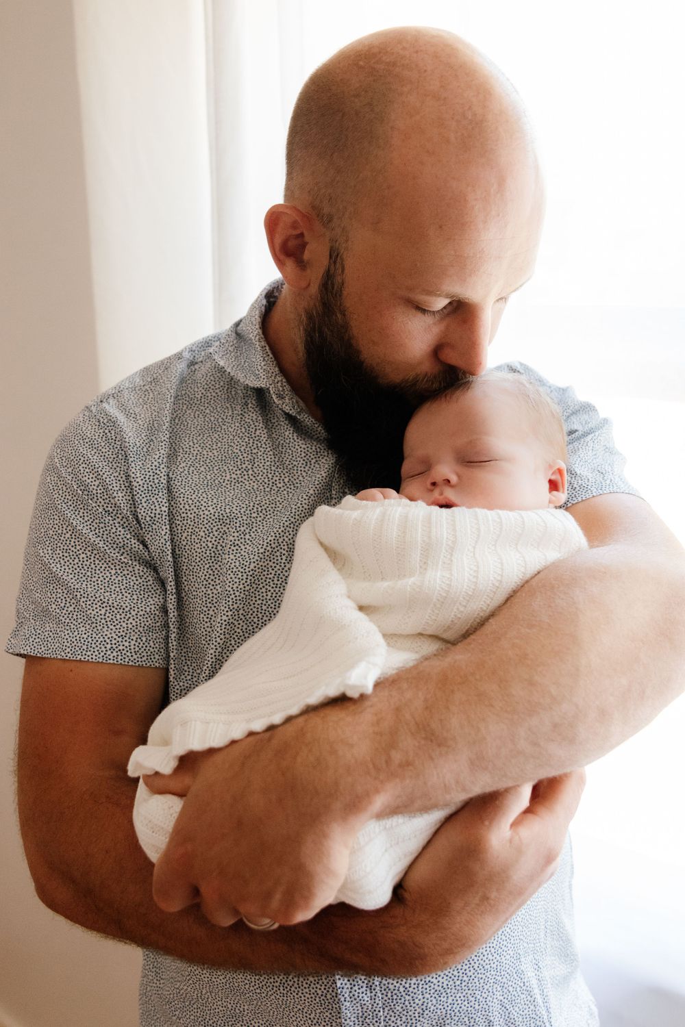 Dad in grey shirt holding and kissing sleeping newborn baby in white outfit. Captured in a natural light studio in the blue mountains Sydney