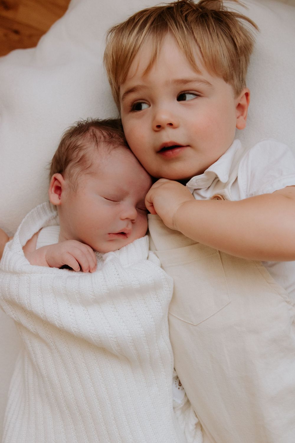 Sweet sibling interaction with newborn baby lying together on white bedding. Captured in a natural light studio in the blue mountains Sydney