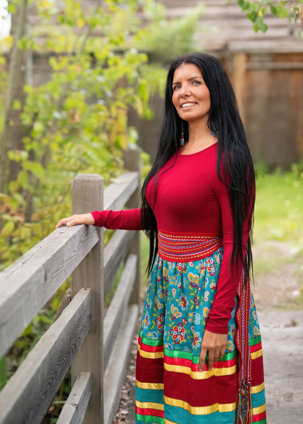 Indigenous woman in red shirt and ribbon skirt on bridge with hand on railing, smiling at camera - Okanagan portrait by Tania Veitch.