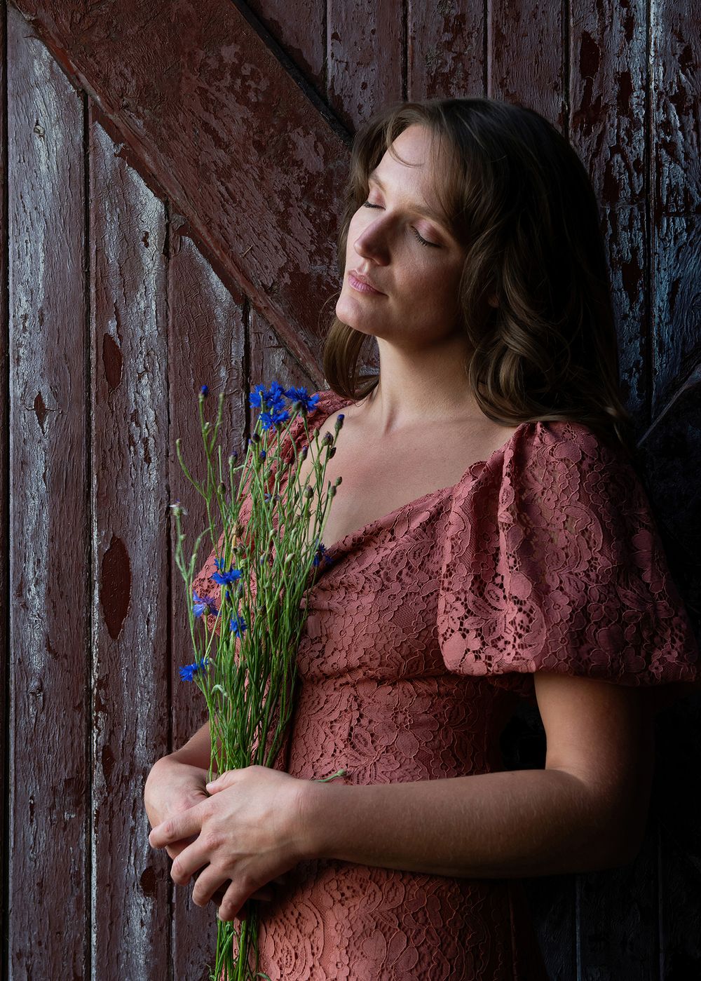 Woman in pink dress holding cornflowers leaning against brown barn door, eyes closed in warm light - Okanagan portrait by Tania Veitch.